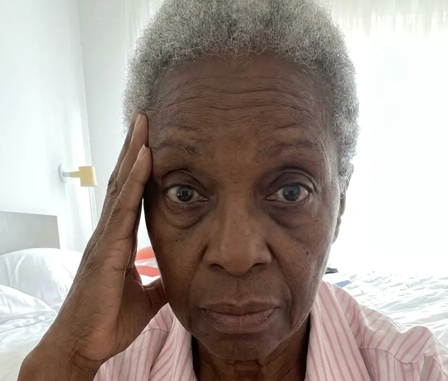 A woman with short grey hair sitting on her bed in striped pyjamas, hand pressed to her temple, looking exhausted and foggy